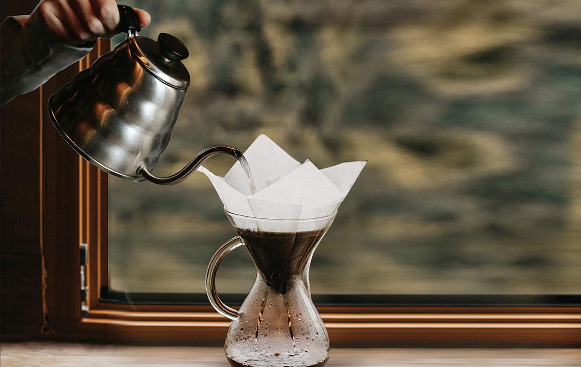 Gooseneck kettle pouring hot water over a paper filter in a glass pour-over carafe, representing Ricco Coffee’s step-by-step brewing guides for making the perfect specialty coffee at home